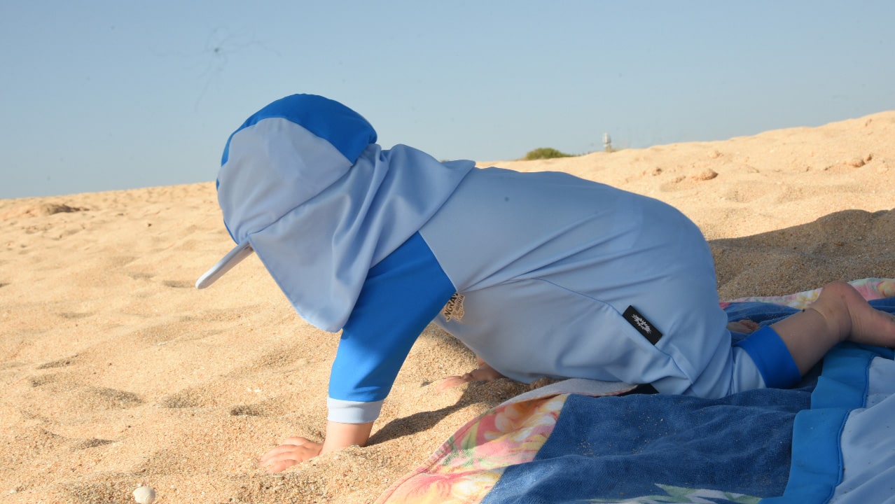 Baby wearing all in one UV suit in blue, rated UPF 50+, on the beach