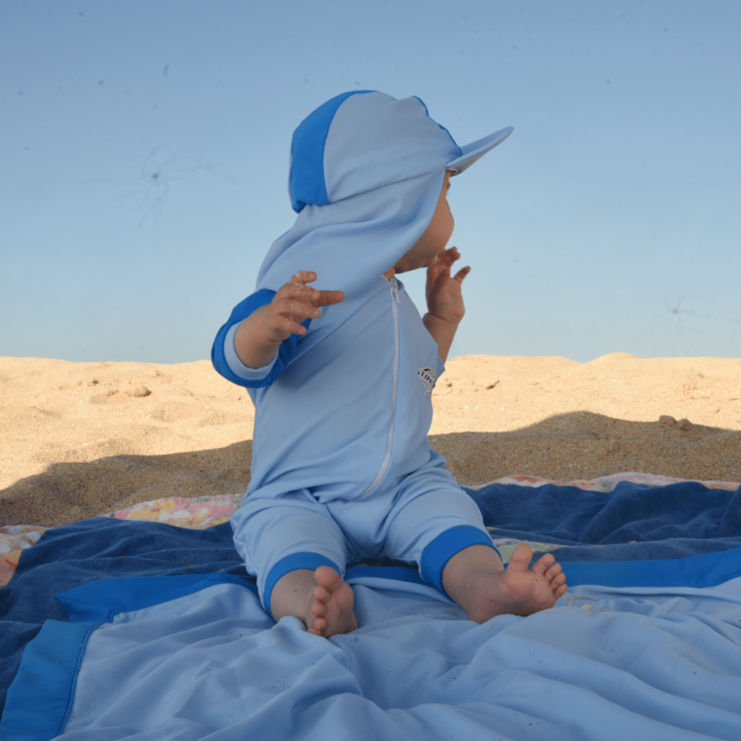 Baby in a blue outfit sitting on a blanket wearing a UPF50+ legionnaire sun hat against a clear sky