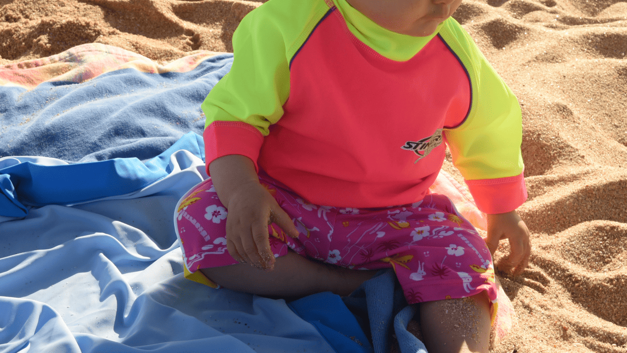 Baby/infant wearing upf swim shorts in colourful design sitting on sand with blue towel