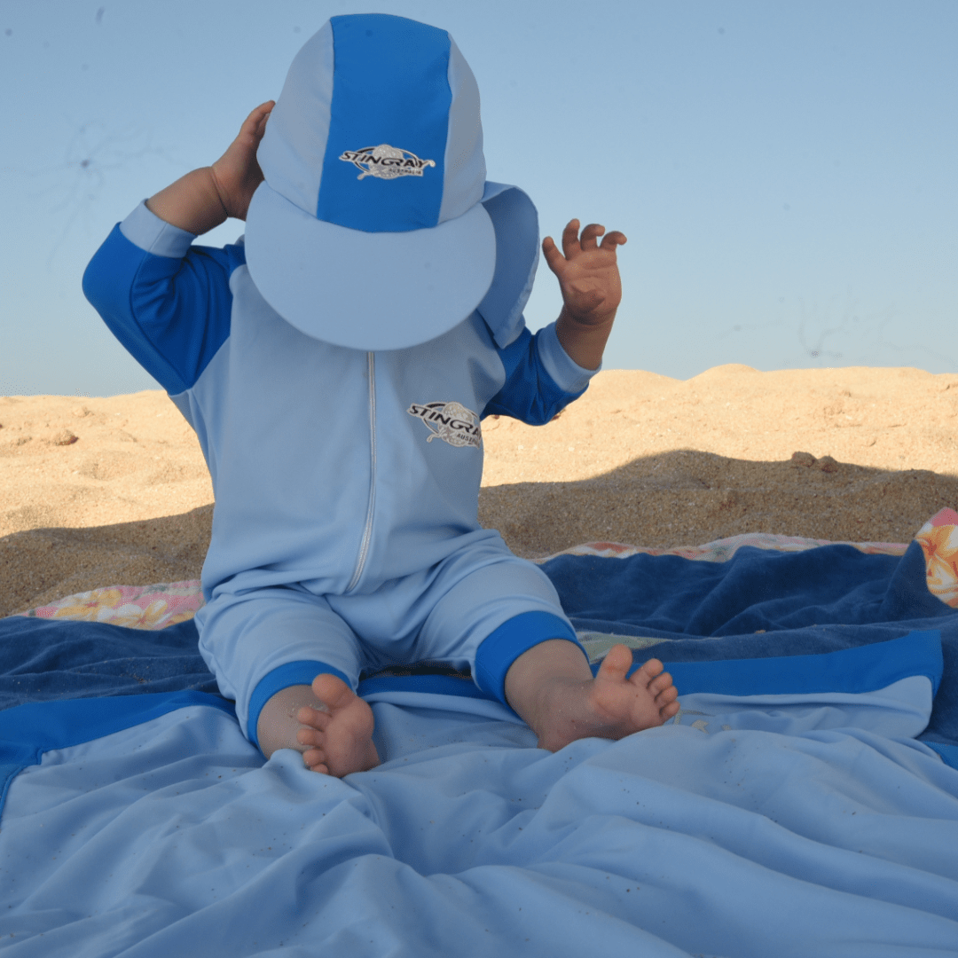 Baby in blue sunsuit and upf cap sitting on a UPF 50+ blanket