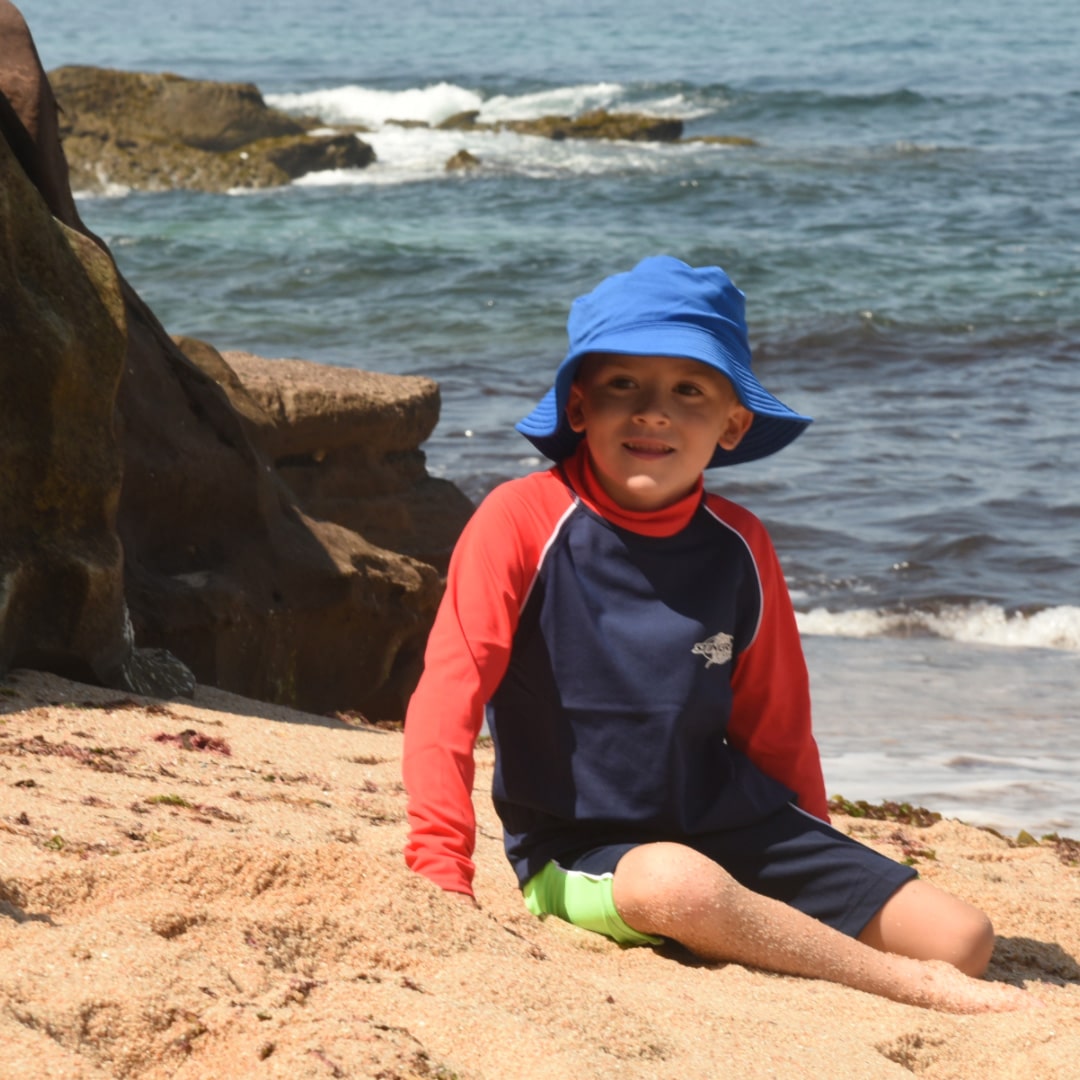 Kiddies swimwear and clothing banner image: child wearing a blue upf sun hat and red and navy upf50 rash shirt sitting on a sandy beach with ocean waves in the background.