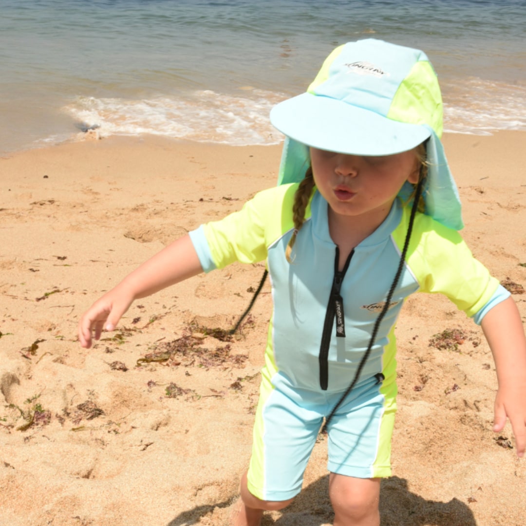 Child in a light blue and yellow UPF 50+ outfit on a sandy beach with ocean waves in the background
