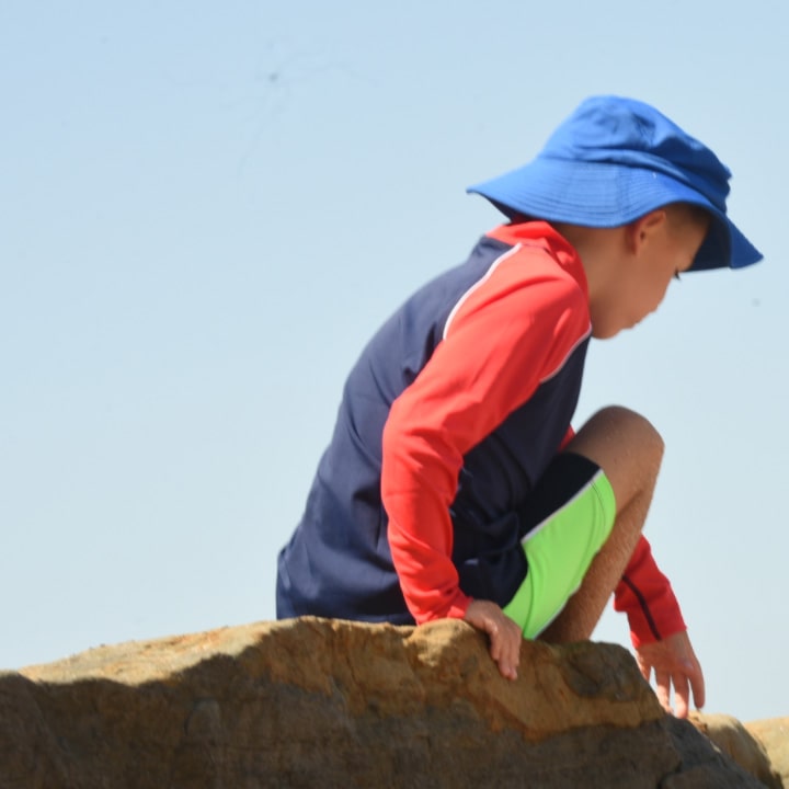 Child wearing uv beachwear including a blue hat and colourful upf rash shirt. He is sitting on a rock against a clear sky.