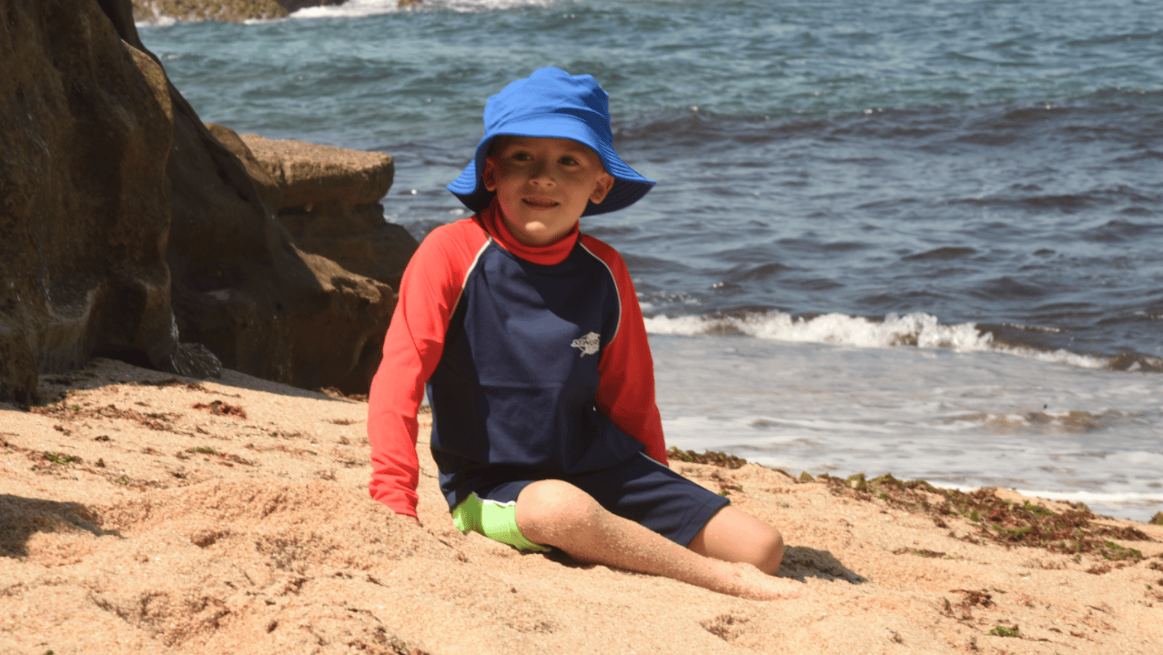Child sitting on a beach wearing a upf blue hat and colourful UPF50 swimwear with ocean waves in the background.
