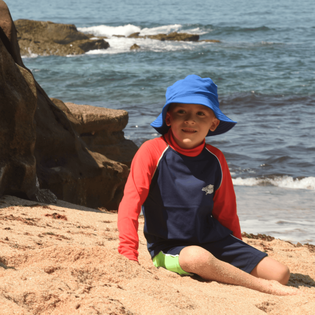 Child wearing a blue upf sun hat and red and navy upf50 rash shirt sitting on a sandy beach with ocean waves in the background.