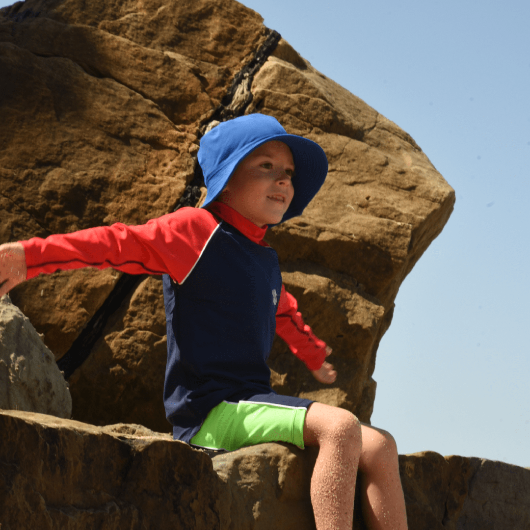 Child wearing a blue UPF 50+ bucket hat and red shirt sitting on large rocks with a clear sky background