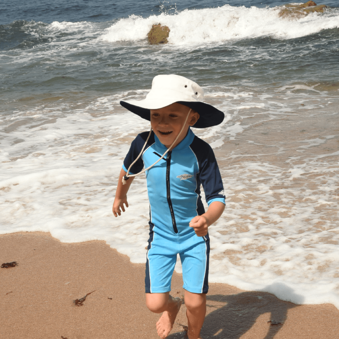 Child in a blue swimsuit and white UPF 50+ sun hat playing on a beach with waves.