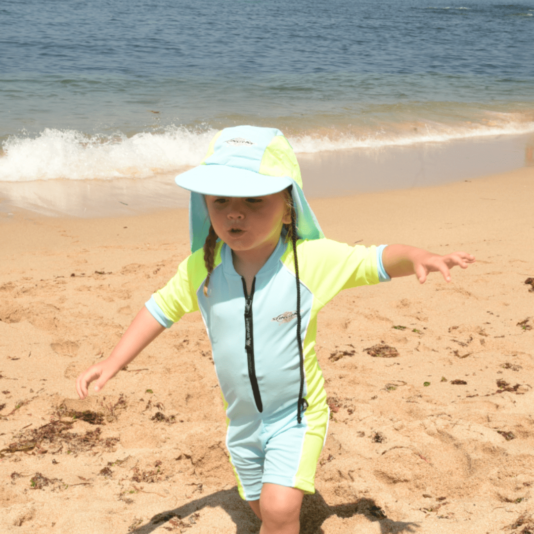 Child in a light blue and yellow upf swimsuit with UPF 50+ legionnaires cap on a sandy beach.