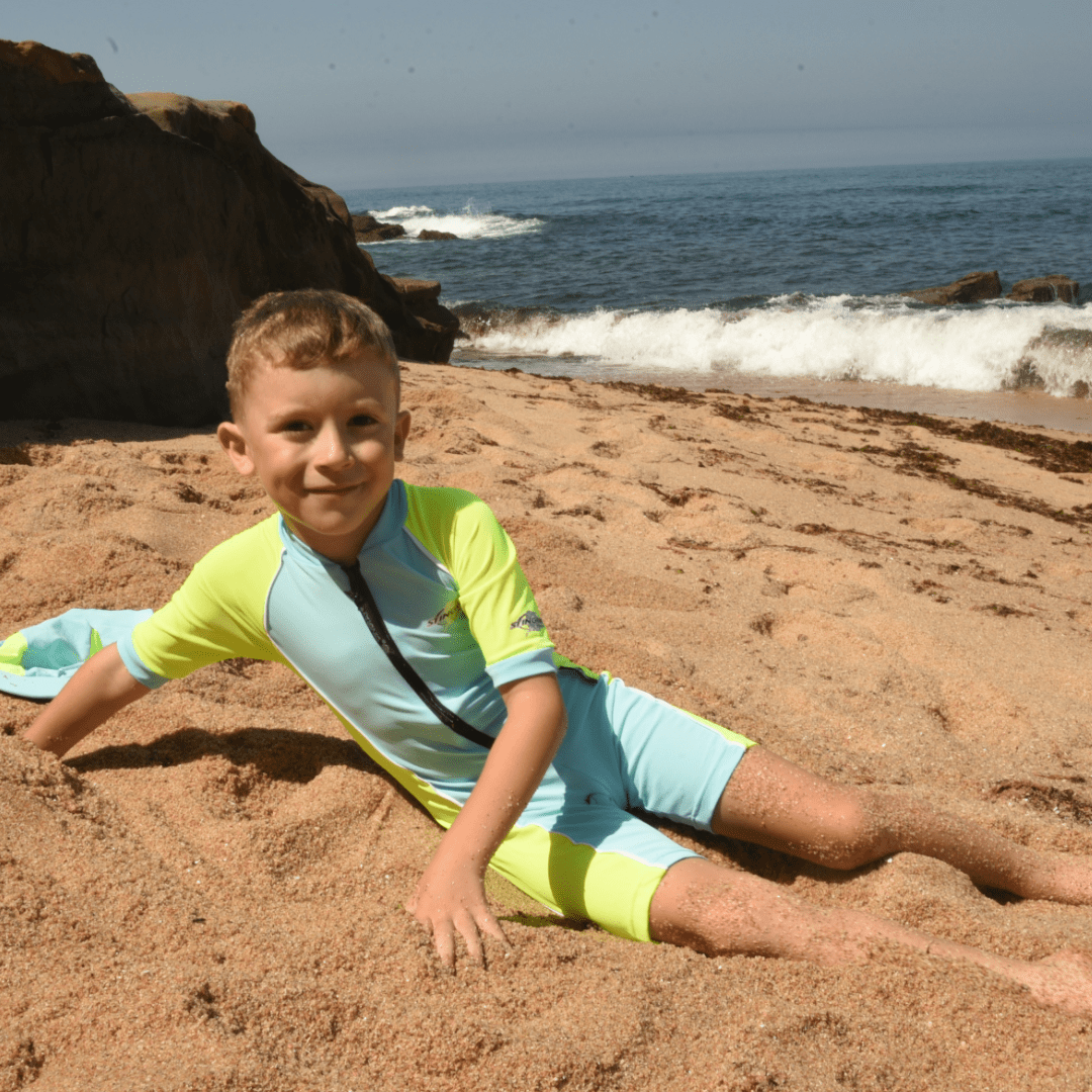Child in a colourful UPF50+ short sleeve swimsuit sitting on a sandy beach with ocean waves in the background