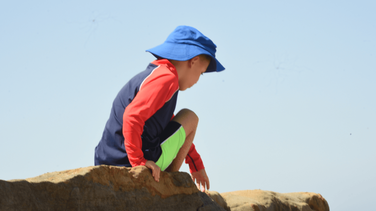 Child wearing a blue hat and colourful upf rash shirt sitting on a rock against a clear sky.
