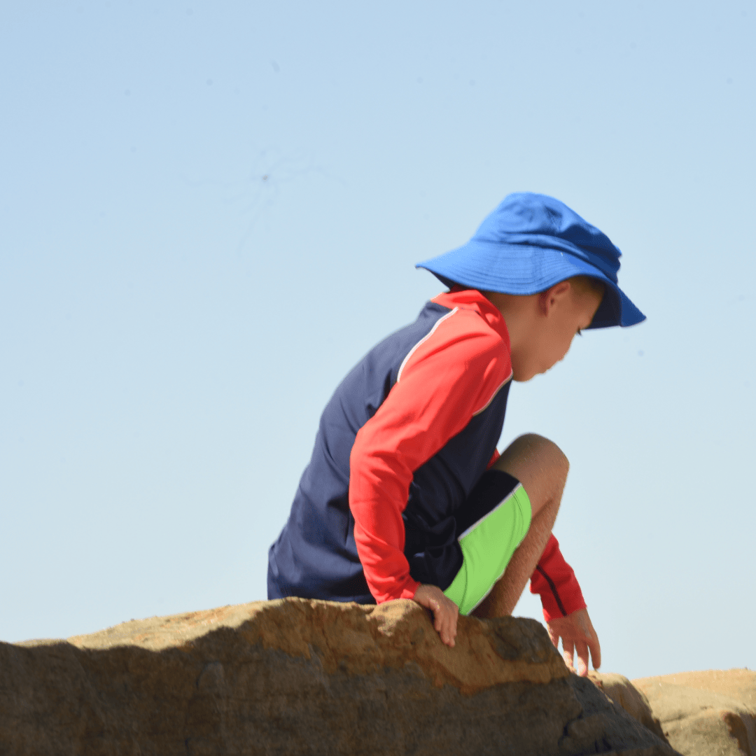 Child wearing a blue hat and upf50 red rash shirt sitting on a rock with a clear blue sky.