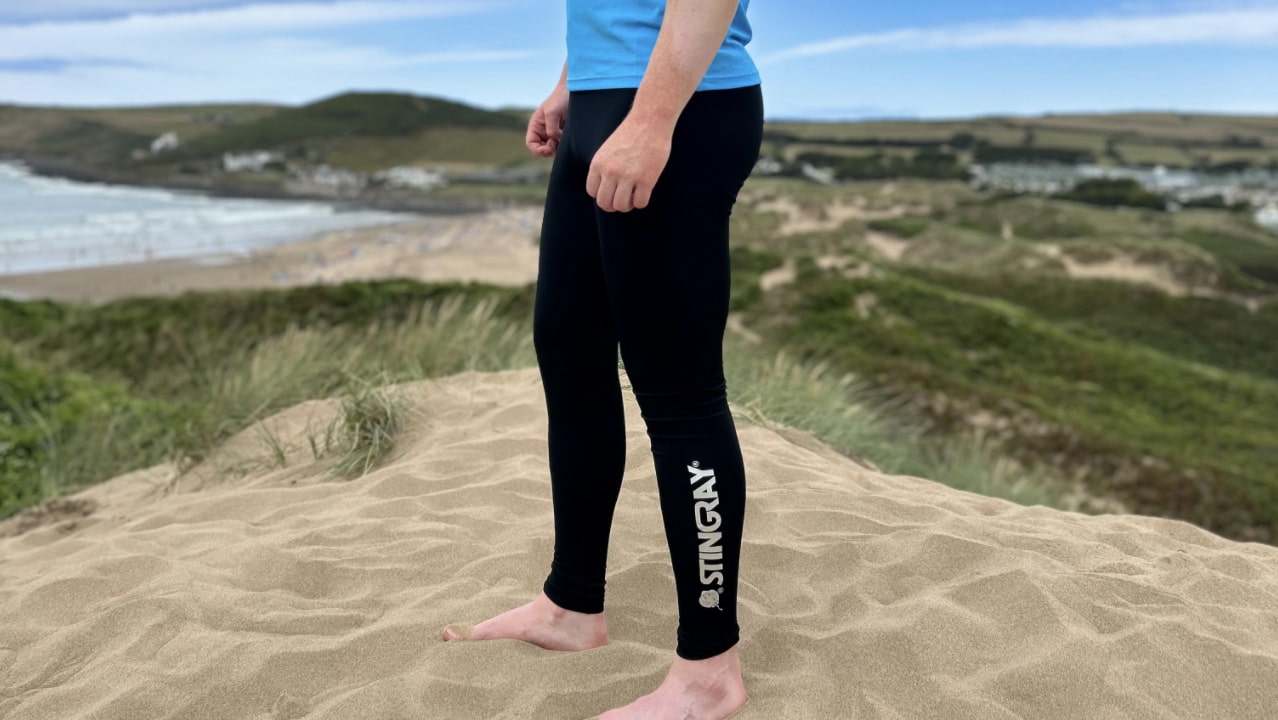 Man wearing black upf leggings with stingray logo, on sand dunes holding a surfboard.