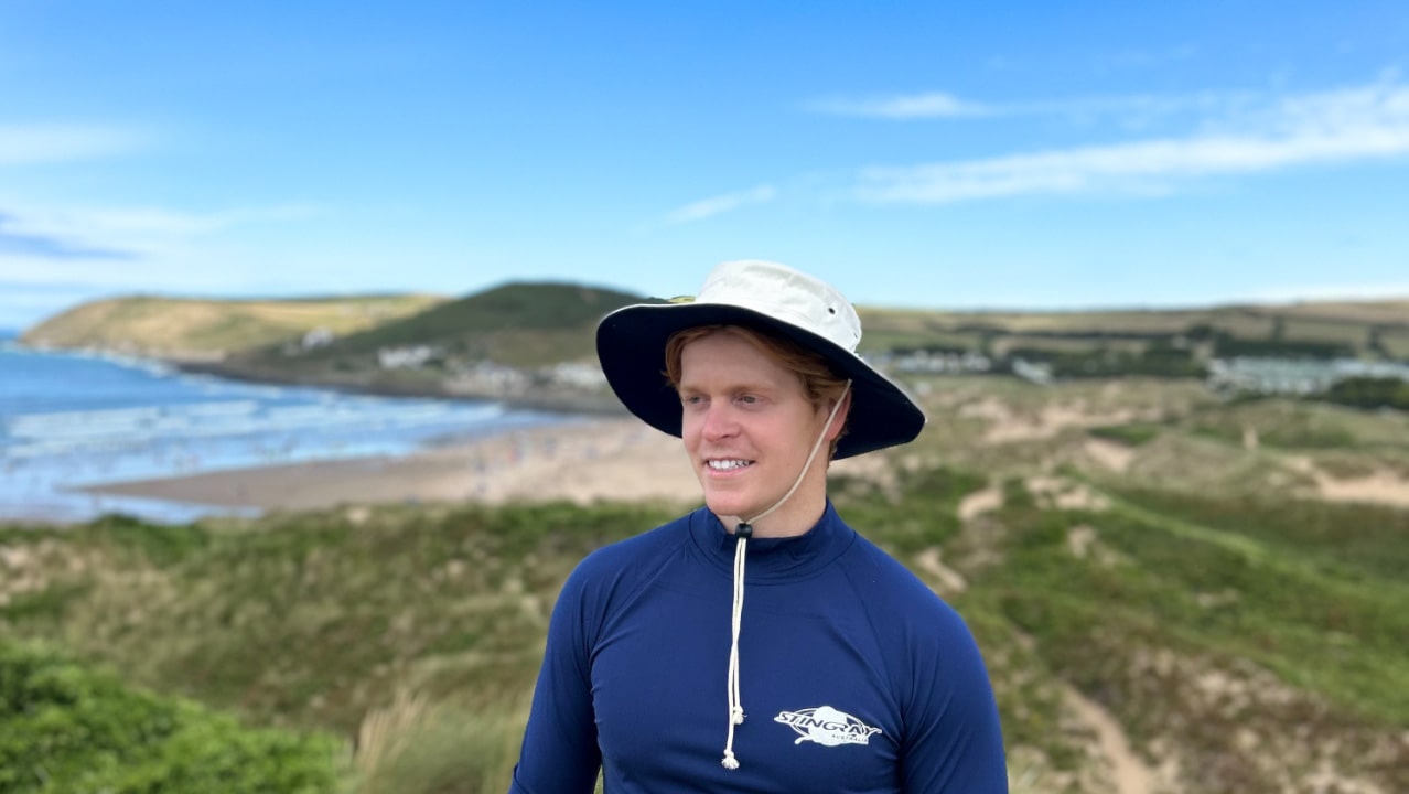 Man wearing a blue upf rash shirt and white hat standing on a coastal landscape with a body of water and hills in the background.