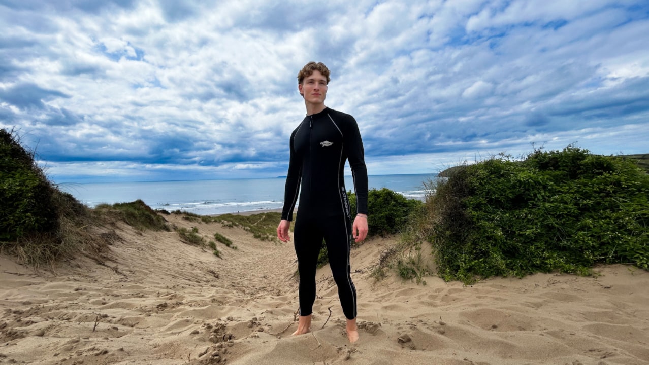Man wearing a black UPF full-body swimsuit standing on a sandy beach with greenery and a cloudy sky.