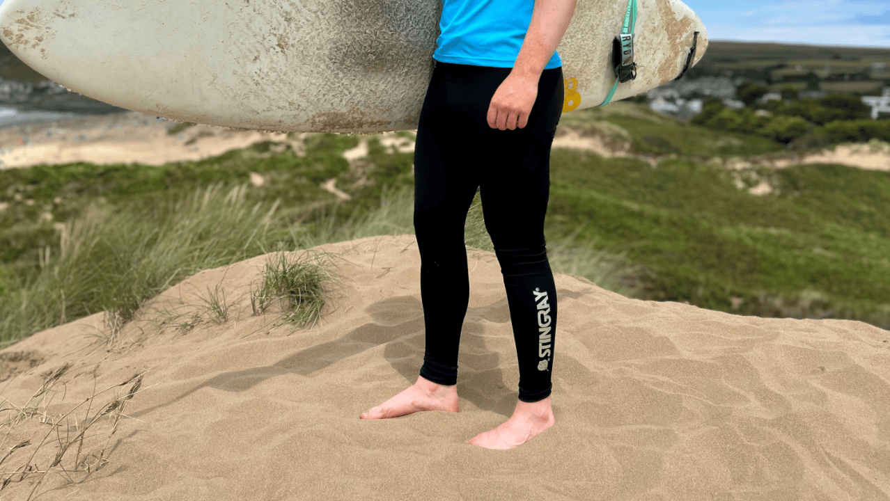 Man wearing black upf leggings with stingray logo, on sand dunes holding a surfboard.