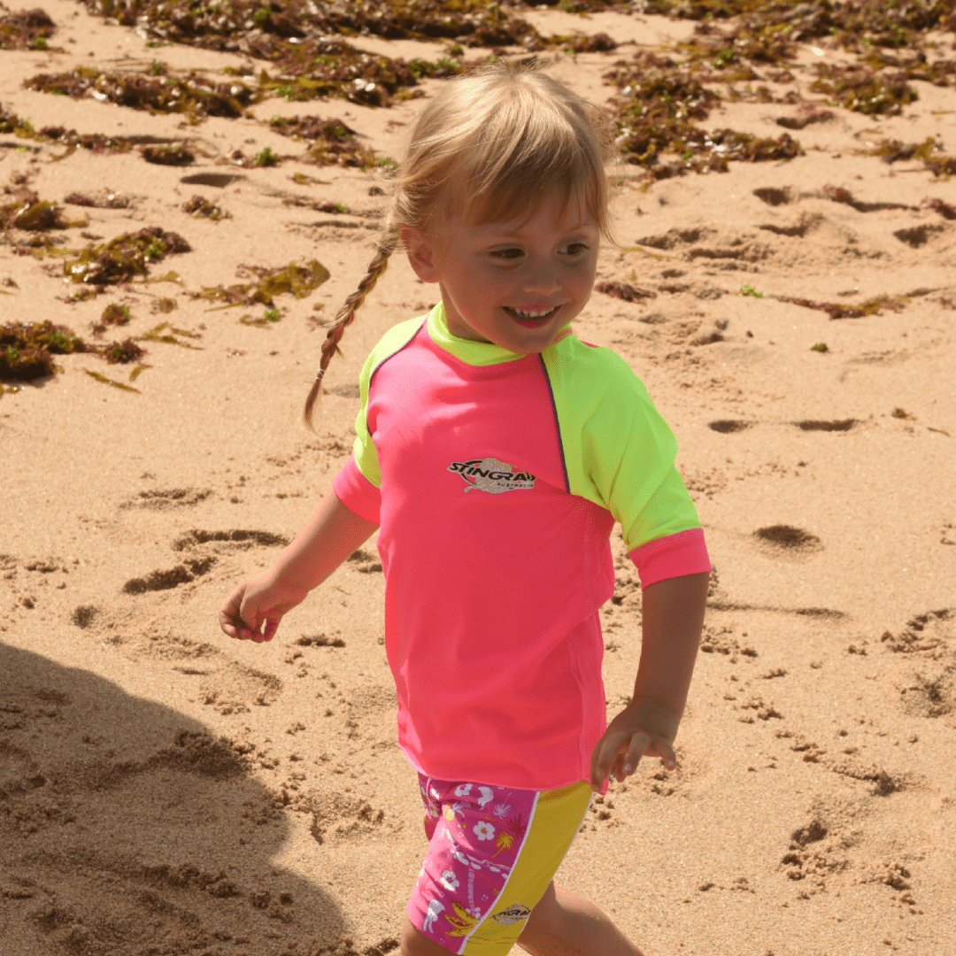 Child wearing a pink and green UPF 50+ rash shirt on a sandy beach
