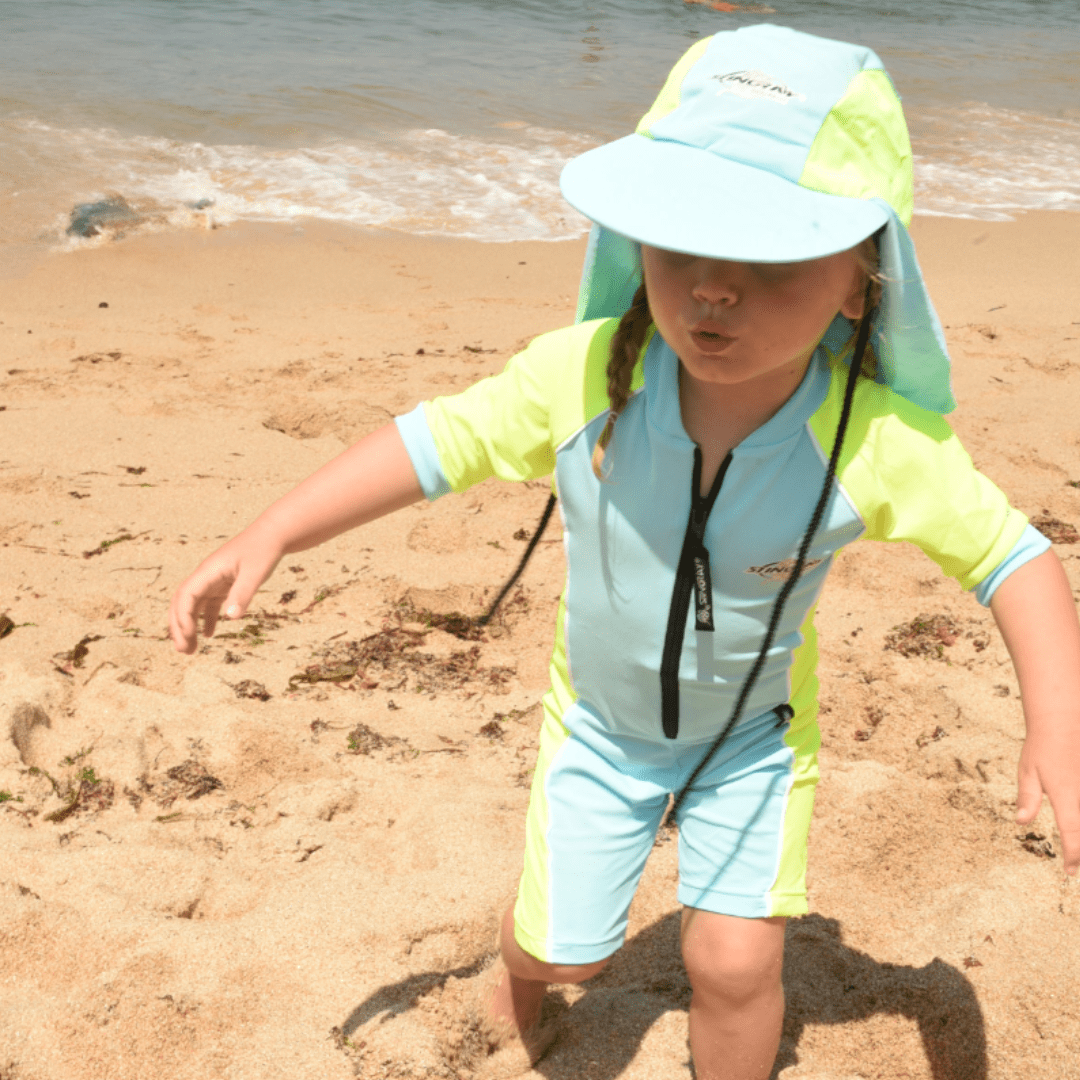 Child wearing a light blue and green sun hat and UPF 50 full body swimsuit on a sandy beach.