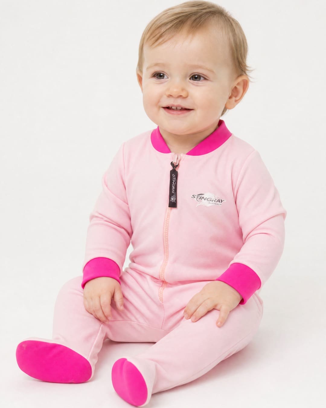 Baby wearing a light pink full-body UV swimsuit/onesie/romper UPF 50+ with dark pink accents on a white background