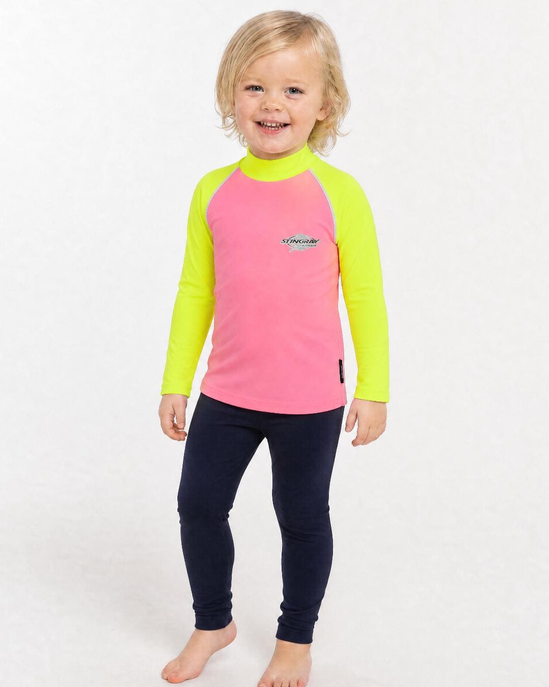 2 year old toddler/child wearing long-sleeve UV rash shirt UPF 50+ in pink and yellow, standing in a white studio