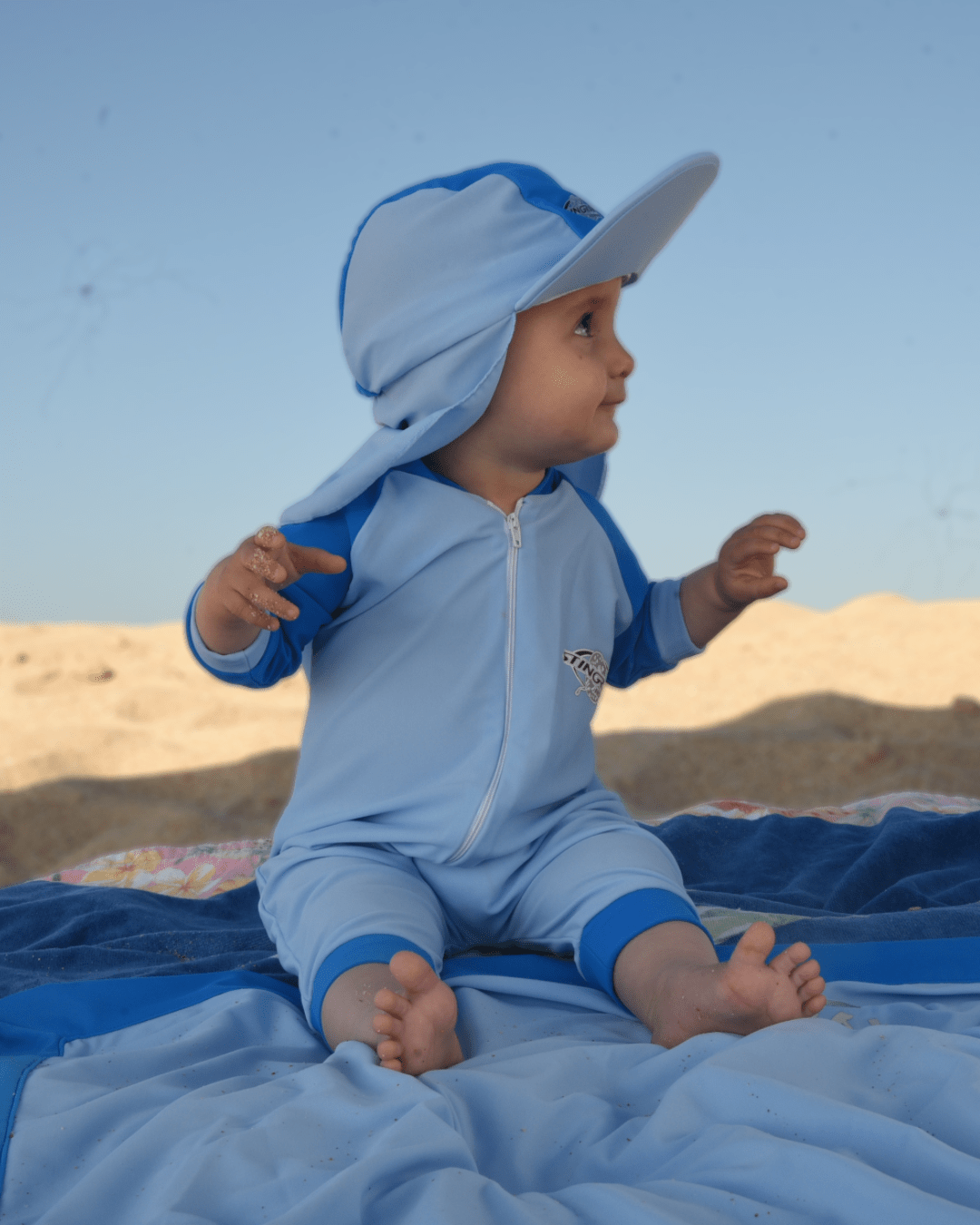 Baby in a blue UV protection swimsuit and hat sitting on a UPF 50+ blanket with a beach landscape in the background