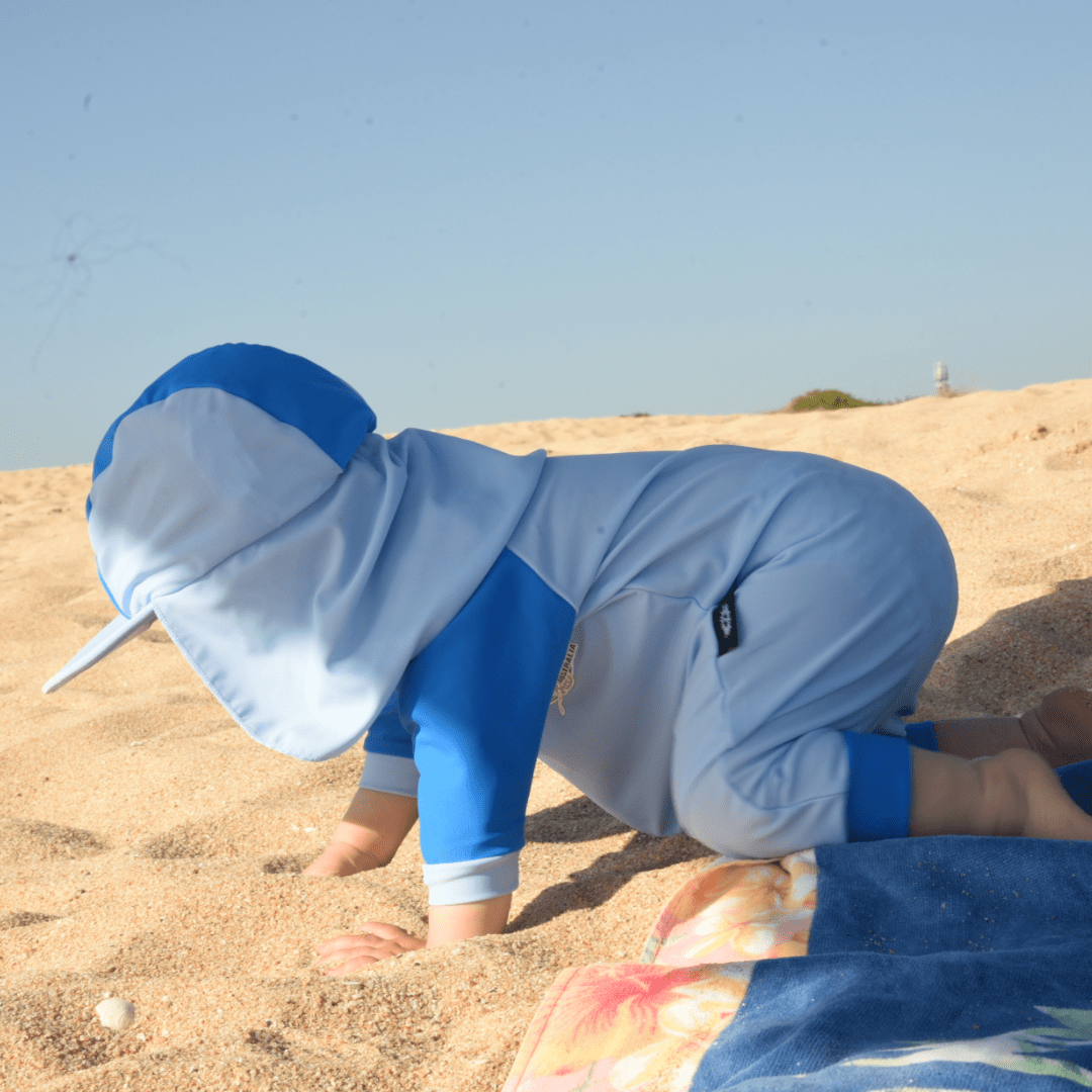 Baby wearing a blue full-body UPF 50+ suit on a sandy beach.