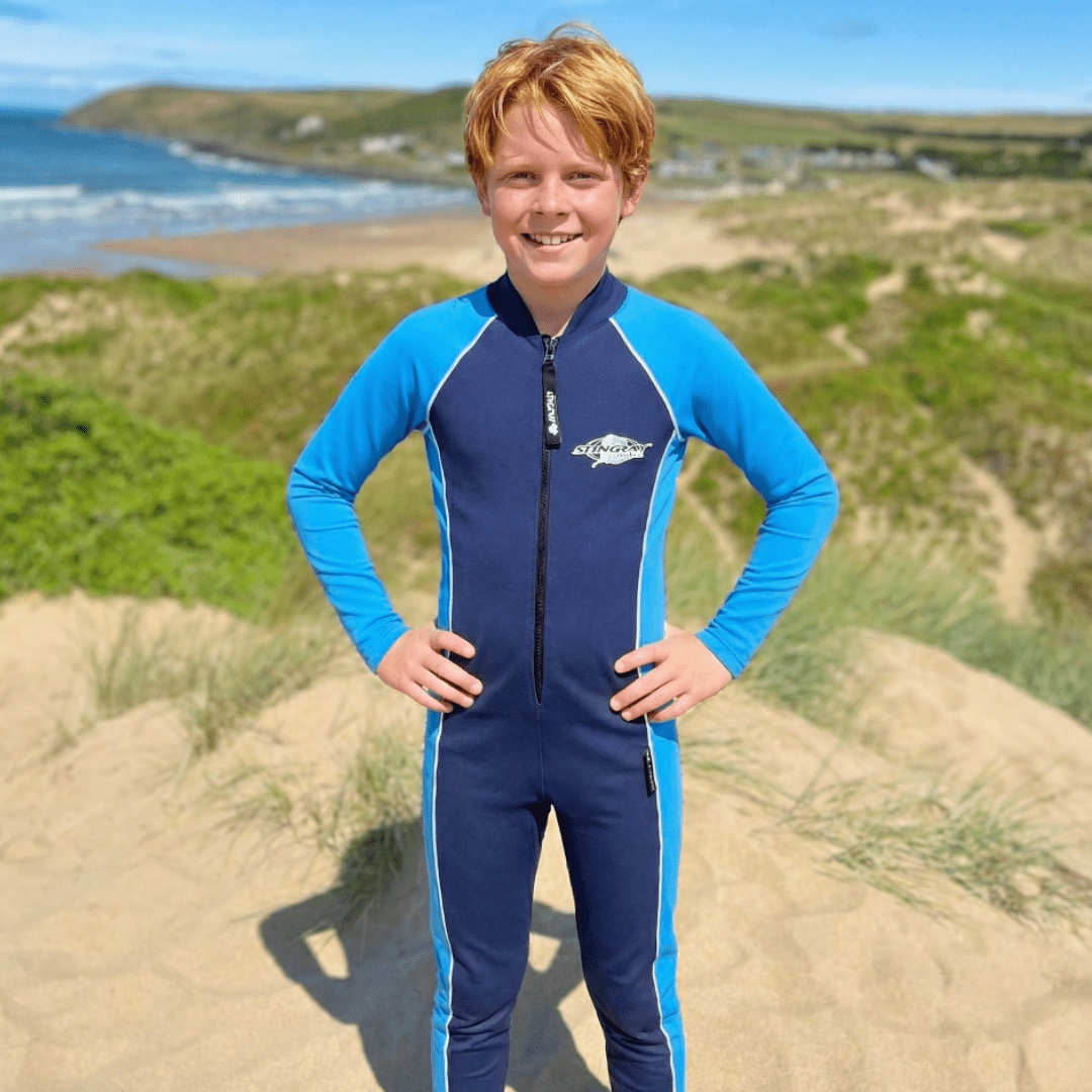 Child wearing a blue and navy full-body UV swimsuit standing on a beach with ocean and grassy dunes in the background.