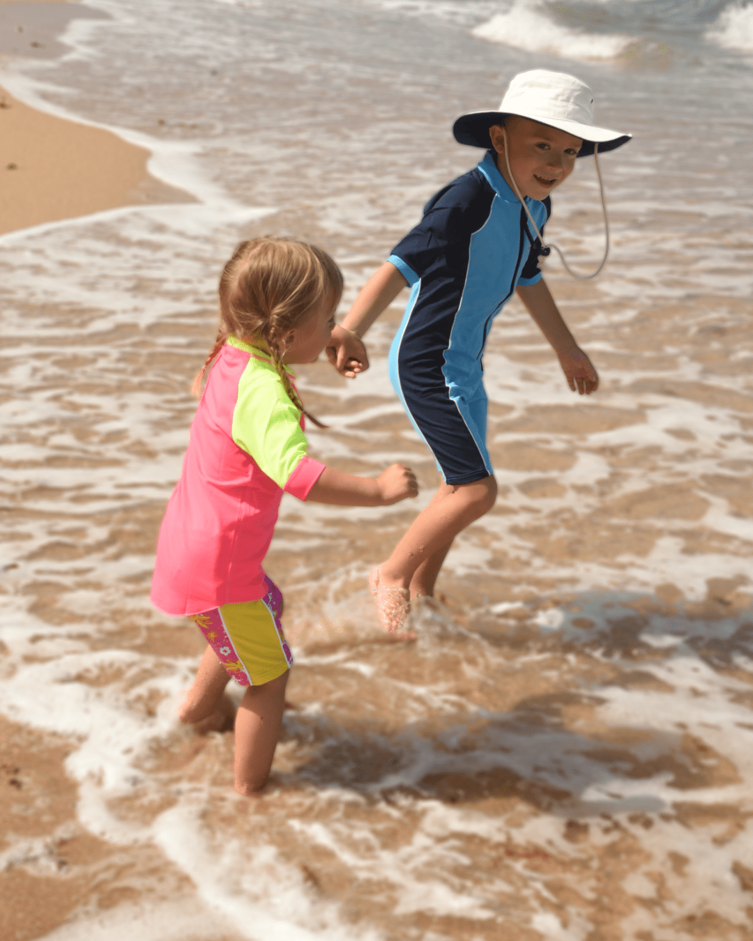 Two children playing in the ocean on a beach wearing UPF 50+ sun protection clothing and swimwear.