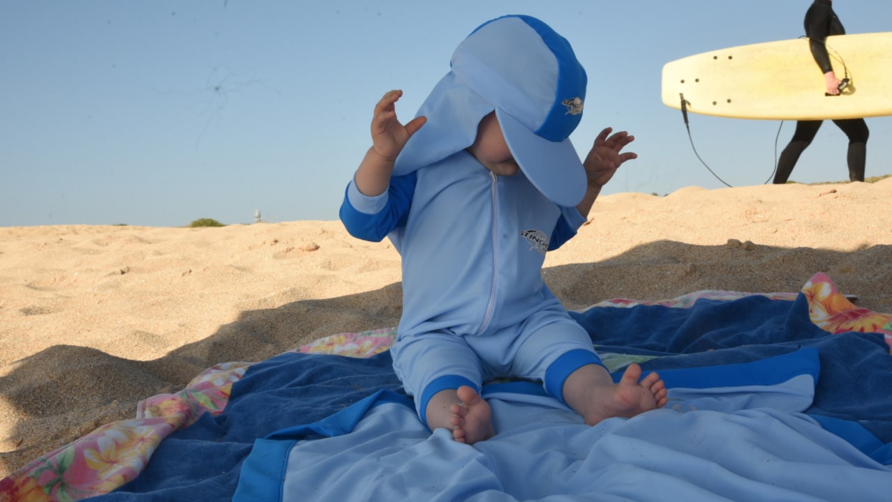 Banner image of a child in blue sun protection outfit and hat sitting on a upf50+ blanket in the sand with a person holding a surfboard in the background, for our infant swimming accessories collection