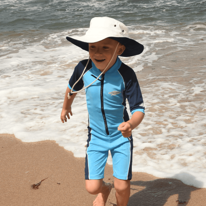 Child in a blue and black UPF swimsuit and UV cricket sun hat playing on a beach.