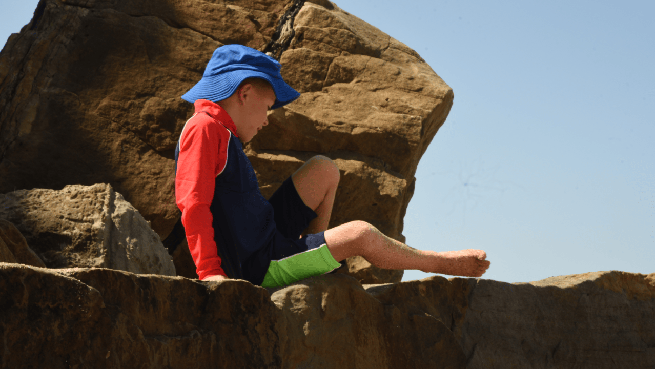 Child sitting on a rock with a blue sun hat and red rash shirt and upf swim shorts, against a clear sky.