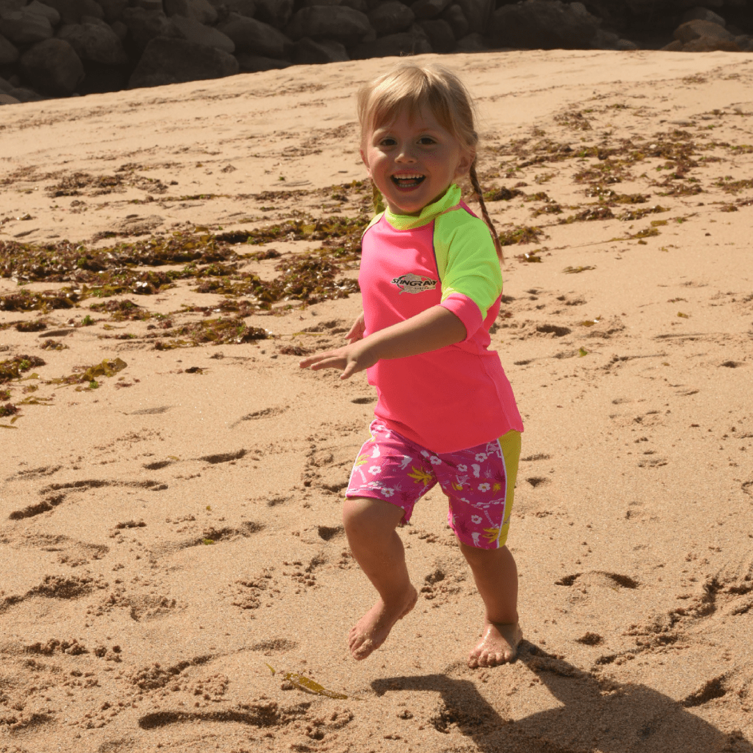 Child in a pink and green shirt and hawaii style UPF 50+ shorts playing on a sandy beach.