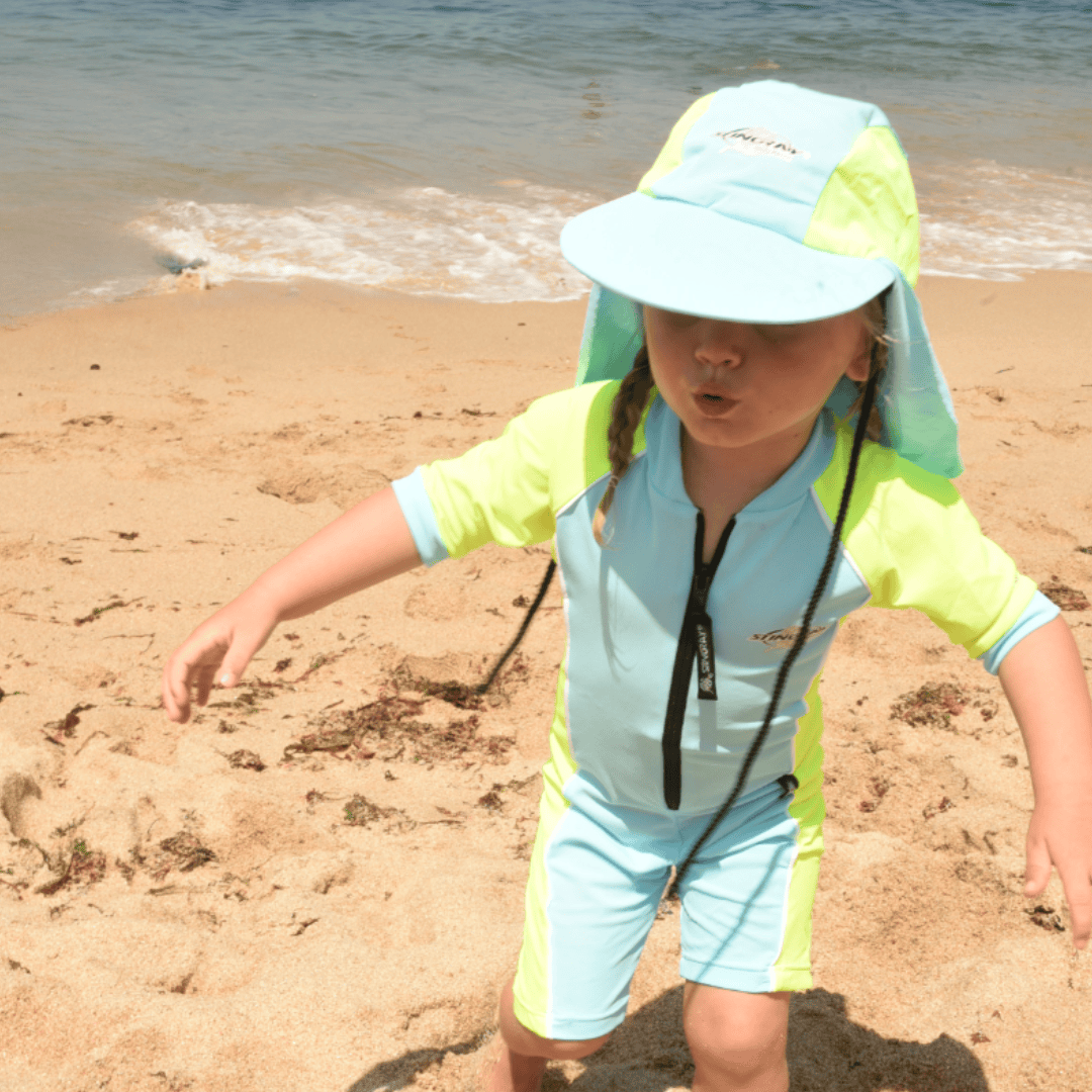 Child wearing a light blue and yellow upf50 sun hat and outfit on a sandy beach.