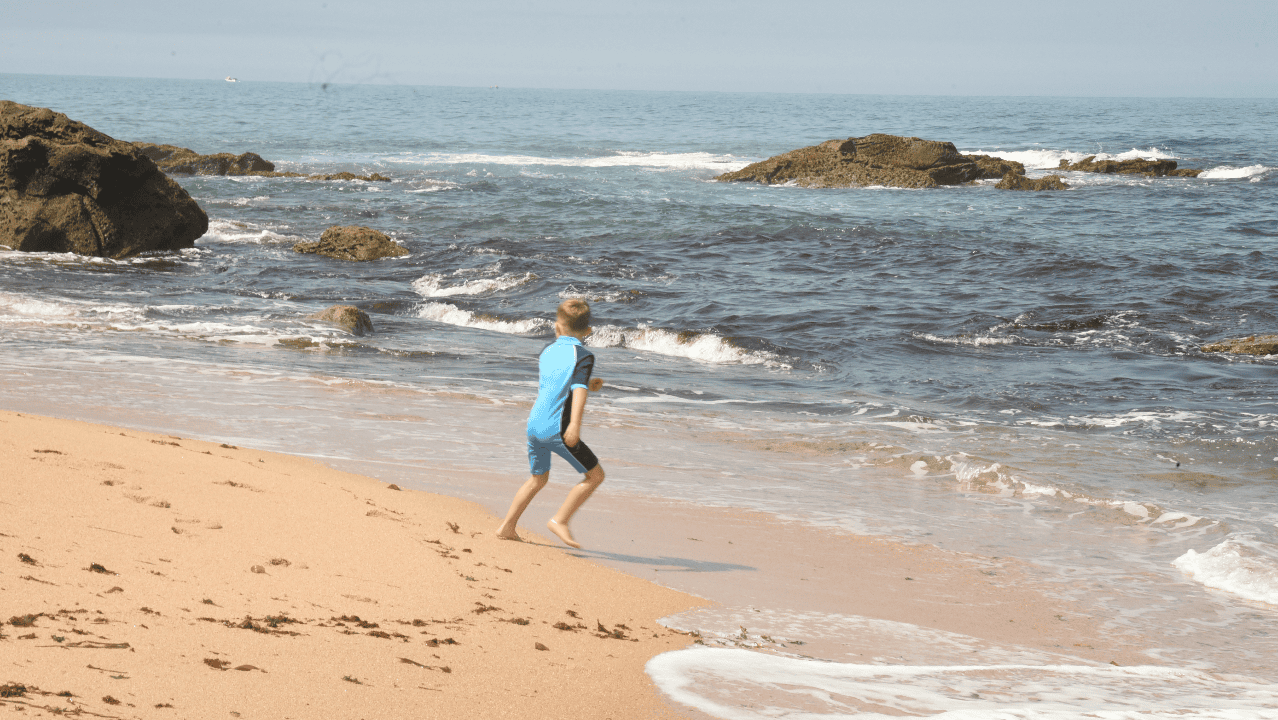 Child wearing upf swimsuit walking on a sandy beach with ocean waves and rocks in the background