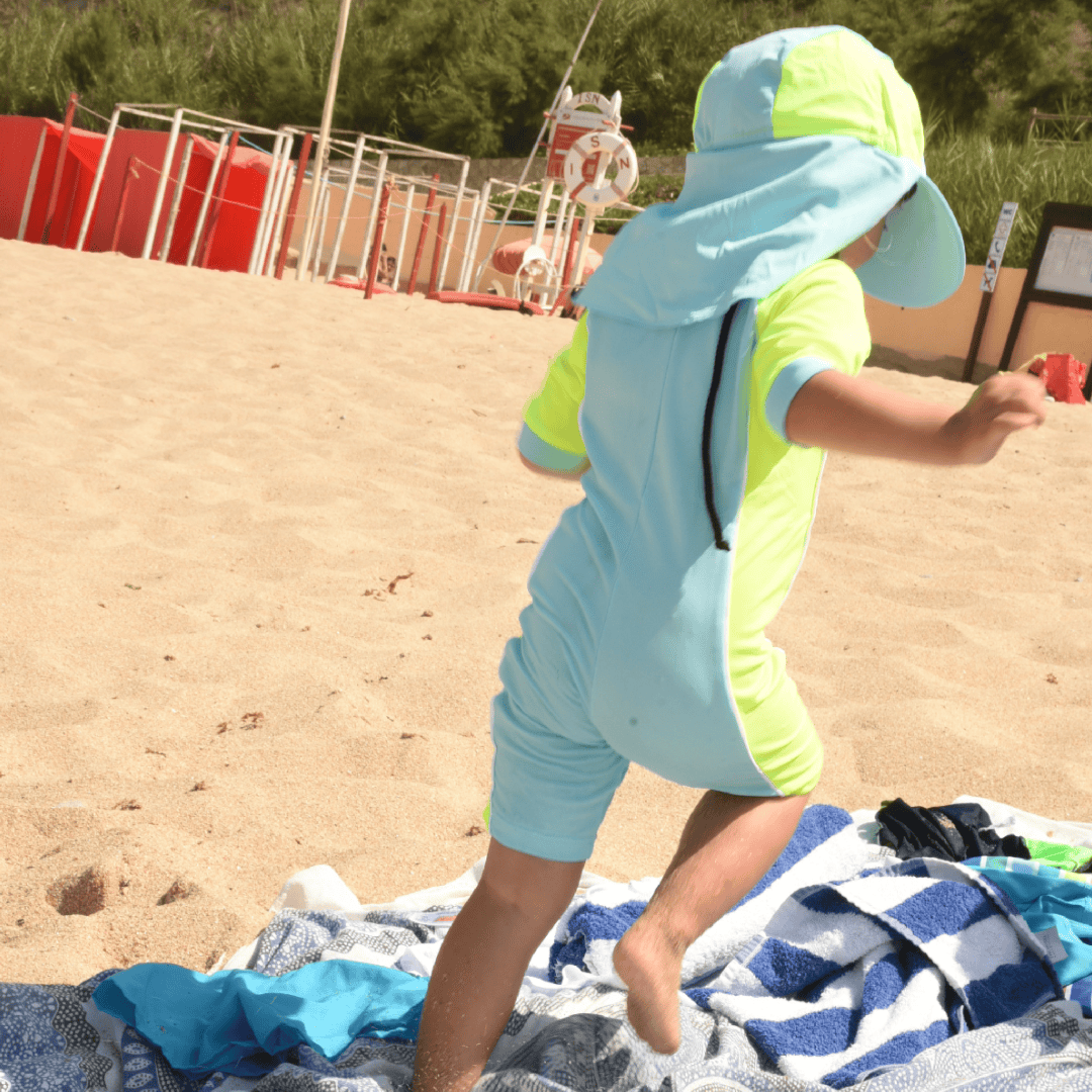 Child in a light blue and green upf swimsuit on a sandy beach with a towel and sand toys.