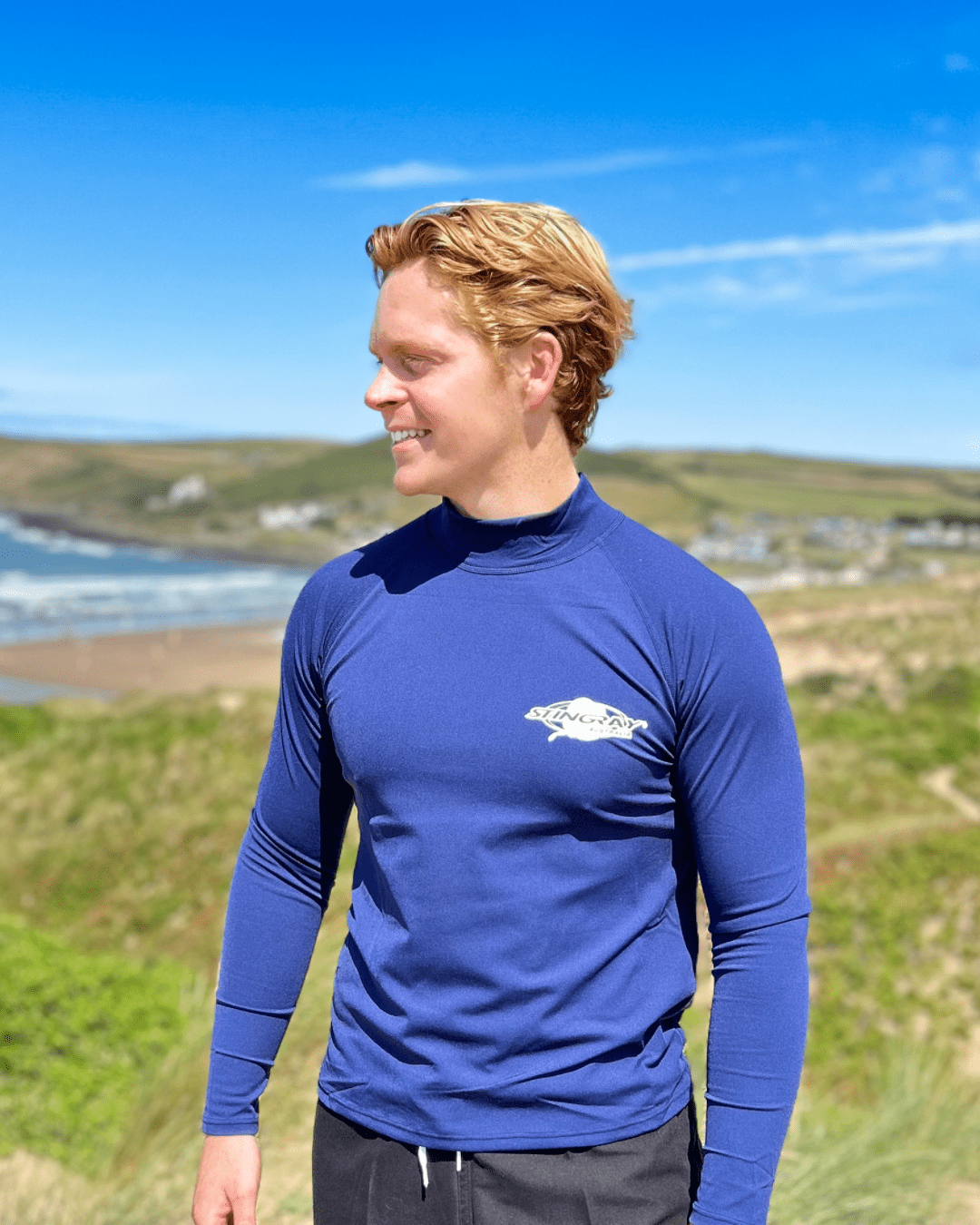 Man wearing a blue long-sleeve UPF 50+ sunsafe shirt with a Stingray logo, standing outdoors with a scenic background.