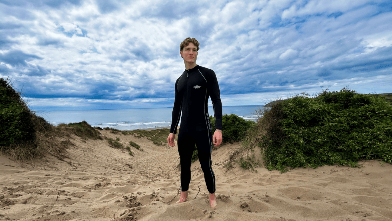 Man wearing a black UPF full-body swimsuit standing on a sandy beach with greenery and a cloudy sky.