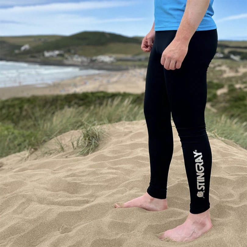 Man wearing black upf leggings with stingray logo, on sand dunes holding a surfboard.