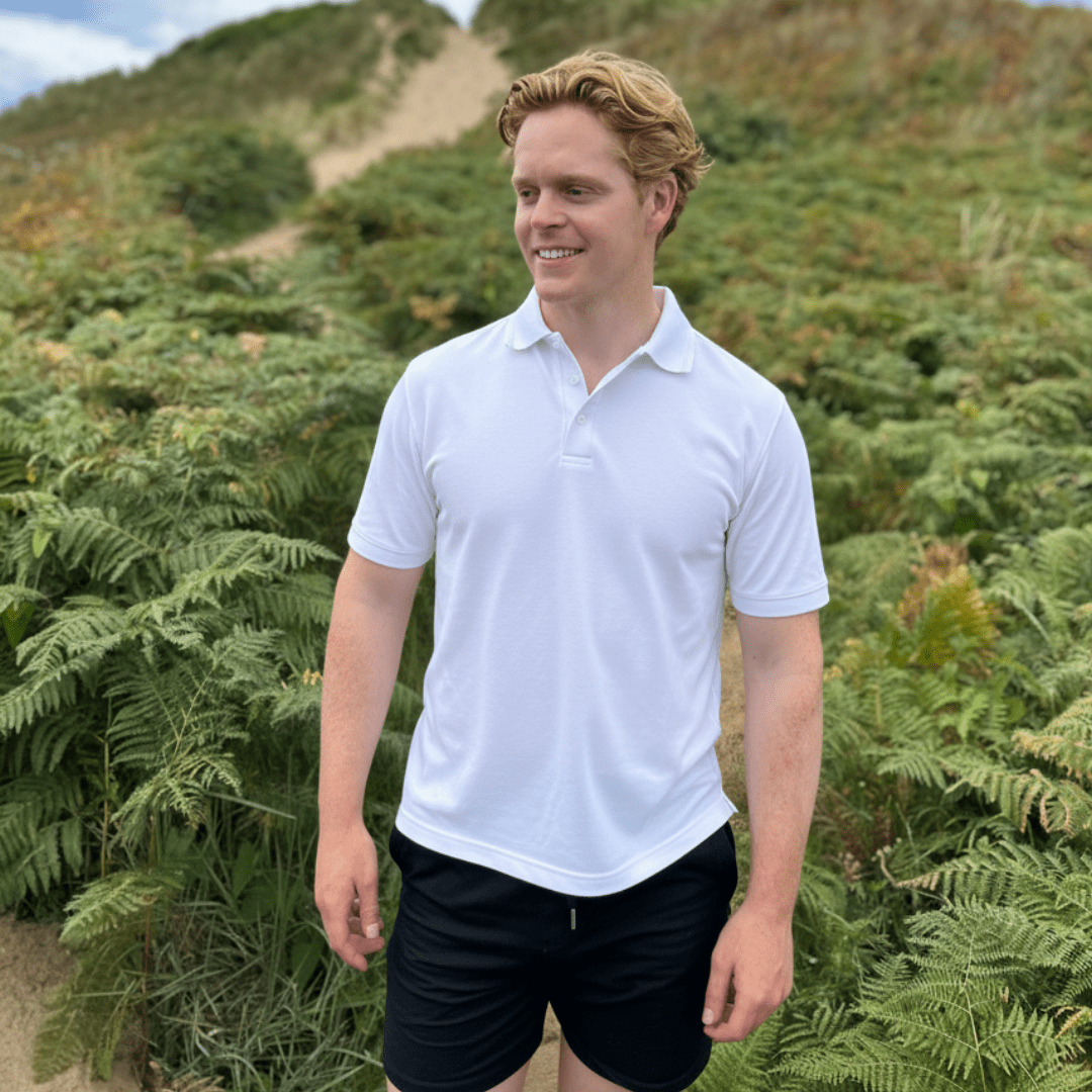 Man wearing a white UV polo shirt and black shorts standing in a natural setting with greenery.