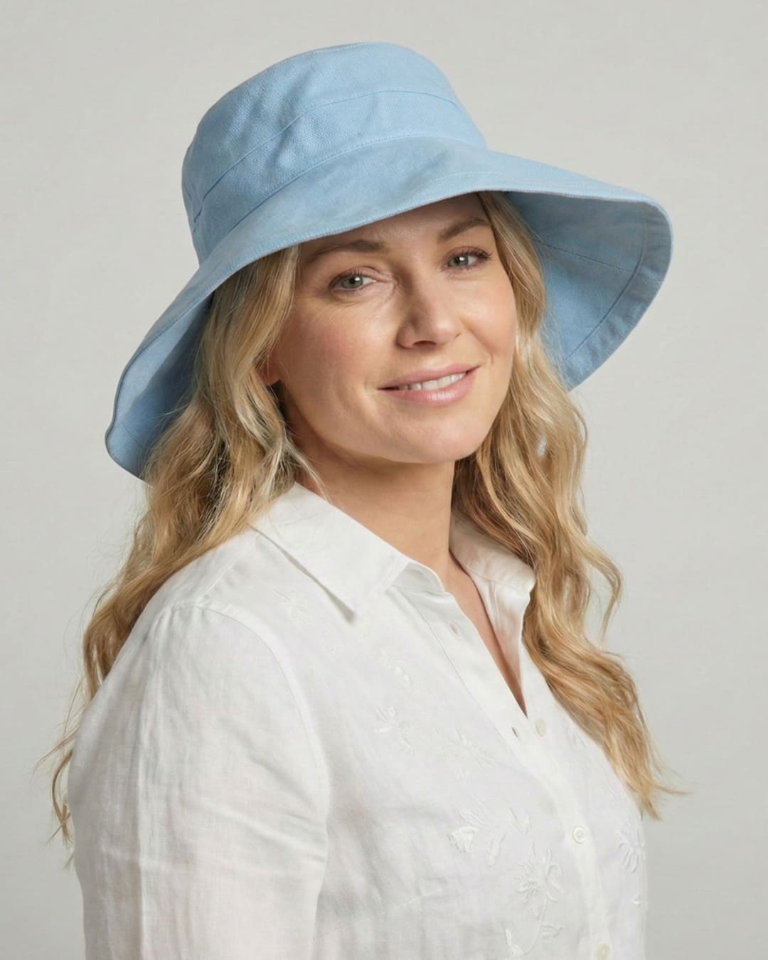 Front-facing 35-year old woman wearing wide brim light blue UPF 50+ sun hat in a grey studio setting