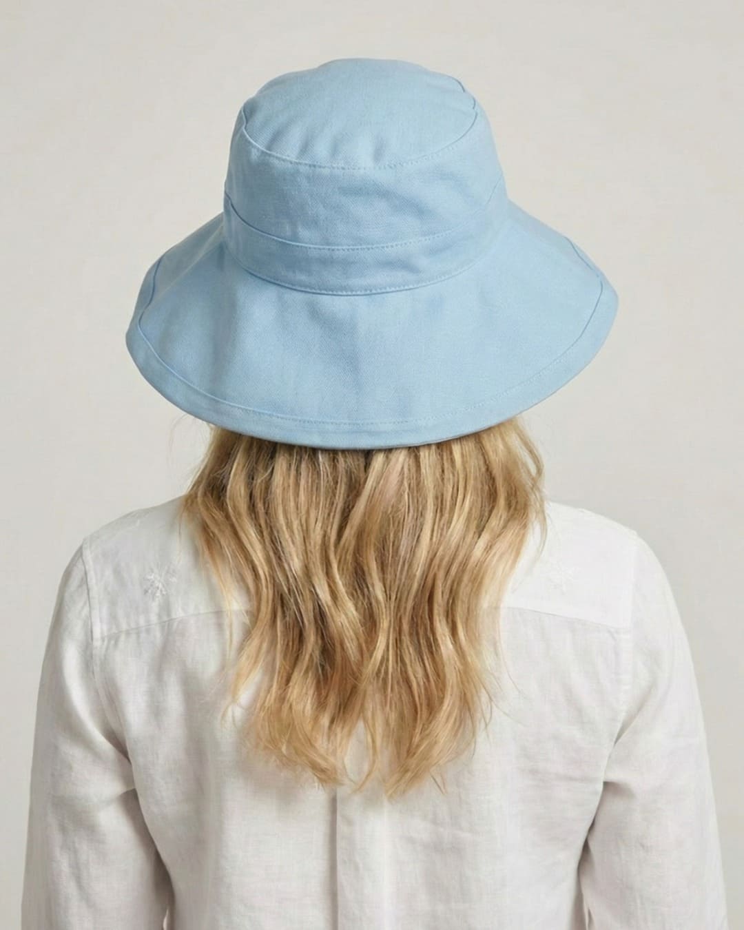 Facing away 35-year old woman wearing wide brim light blue UPF 50+ sun hat in a grey studio setting