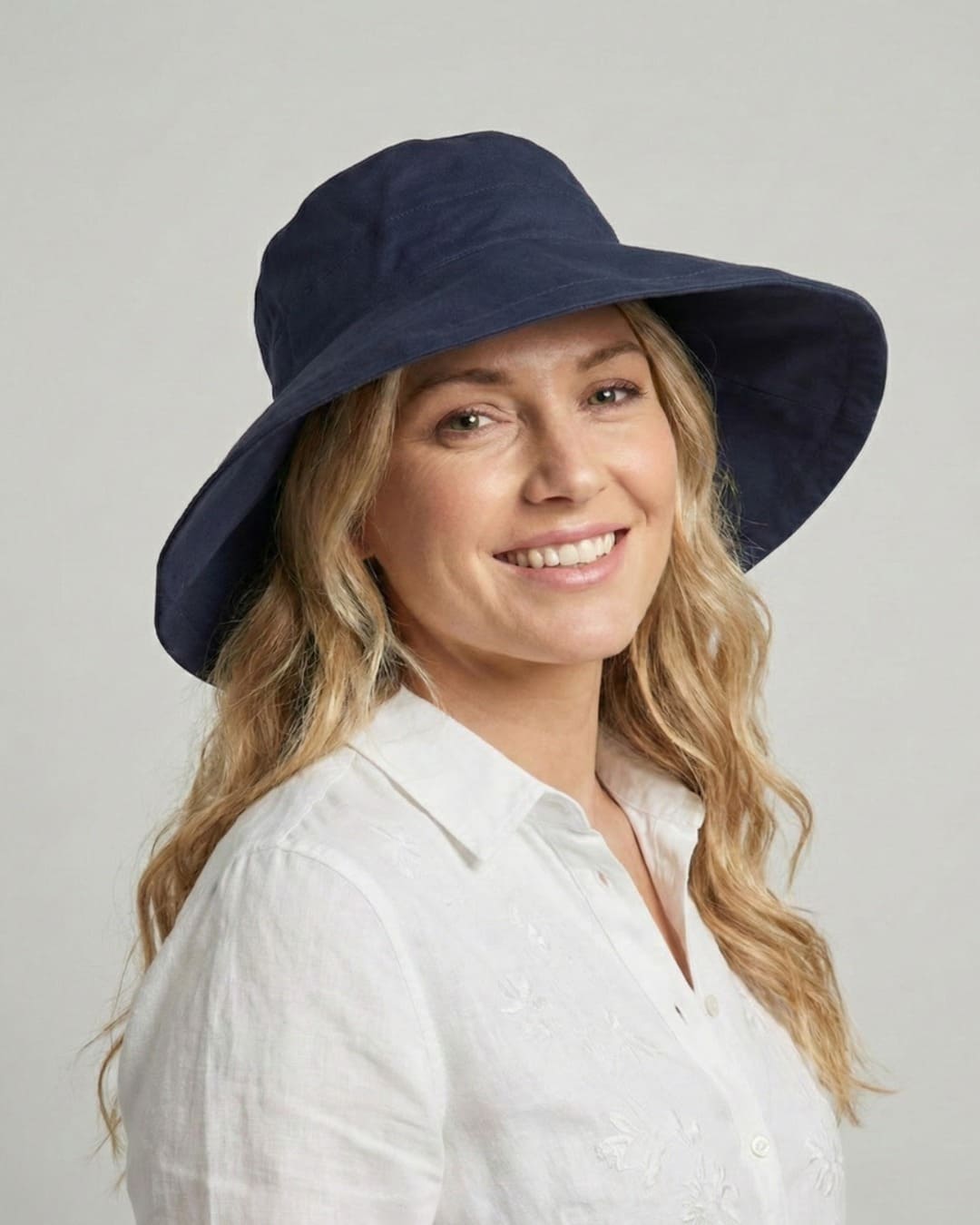 Front-facing 35-year old woman wearing wide brim navy UPF 50+ sun hat in a grey studio setting