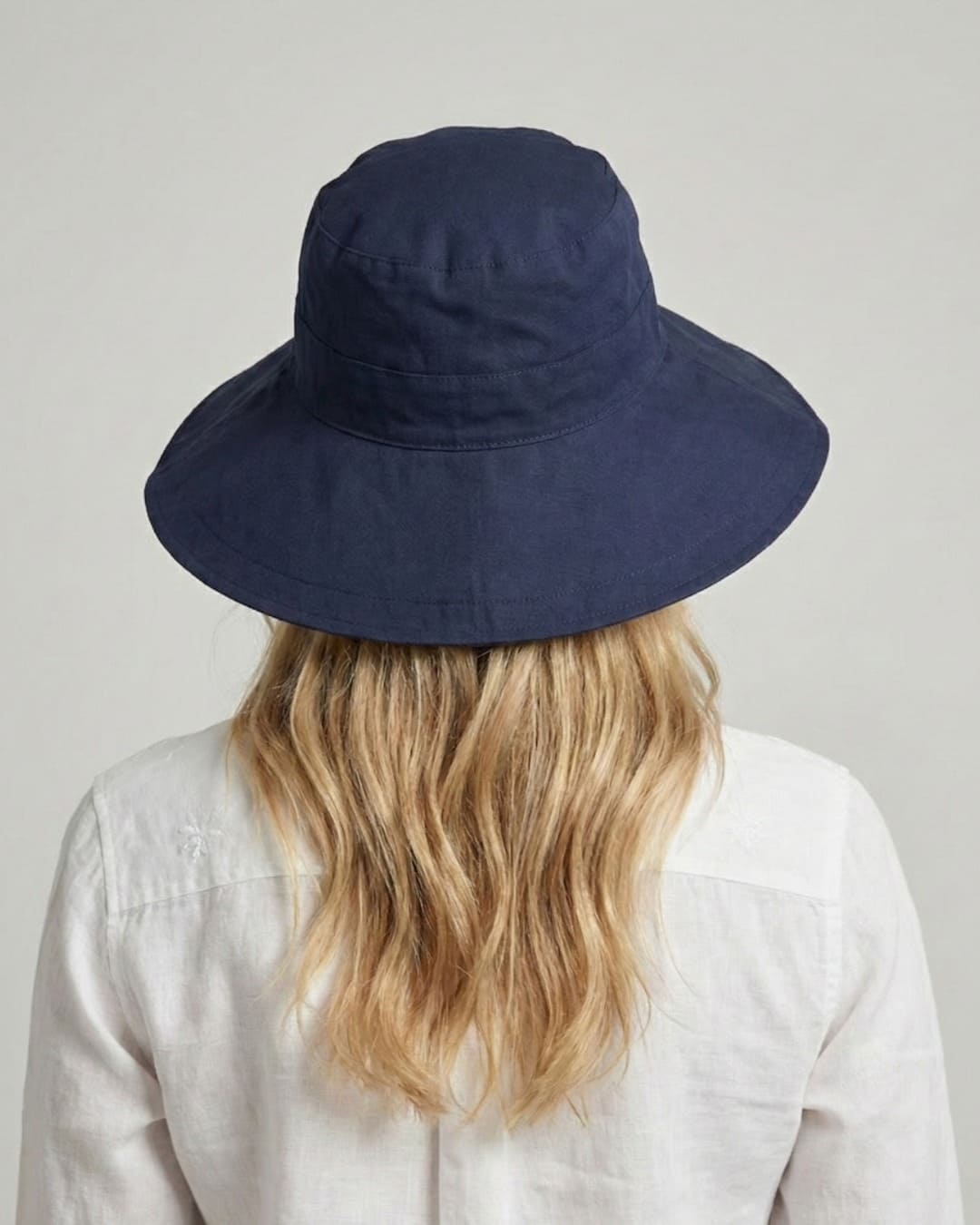 Facing-away 35-year old woman wearing wide brim navy UPF 50+ sun hat in a grey studio setting