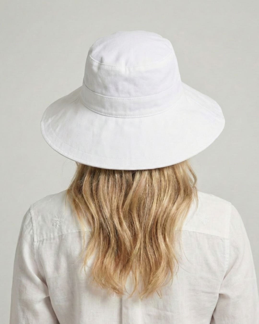 Facing-away 35-year old woman wearing wide brim white UPF 50+ sun hat in a grey studio setting