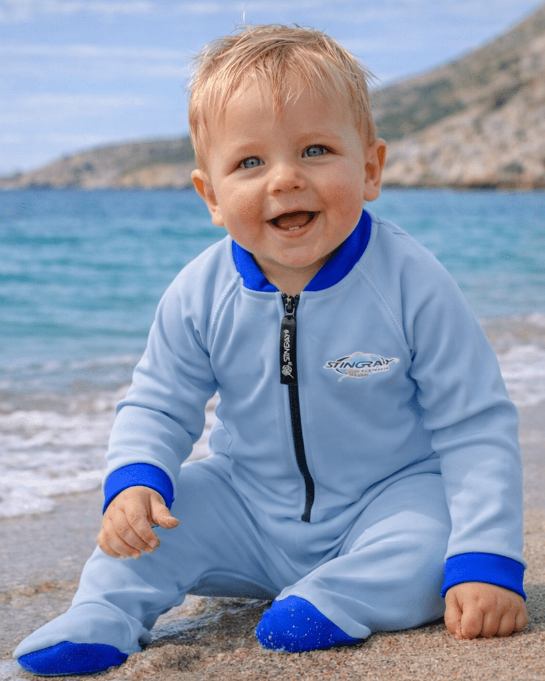 Child wearing a light blue and navy full-body UV swimsuit with 'Stingray' branding on a beach.