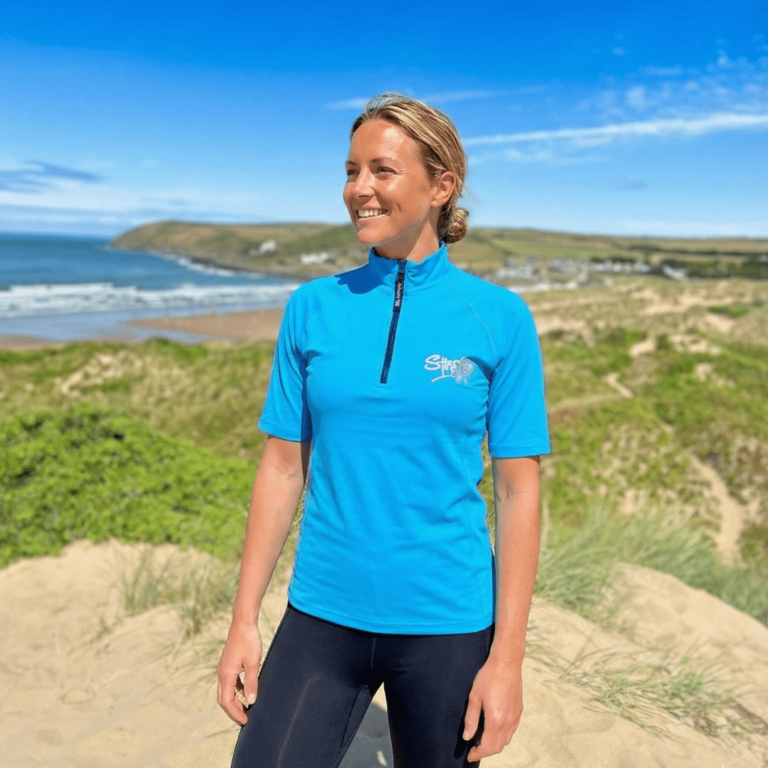 Woman wearing an azure blue UV zip swim top standing on a sandy beach with a scenic background.