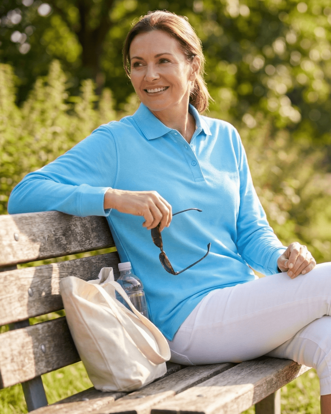 Woman sitting on a bench outdoors wearing a blue long sleeve UV polo shirt and white trousers
