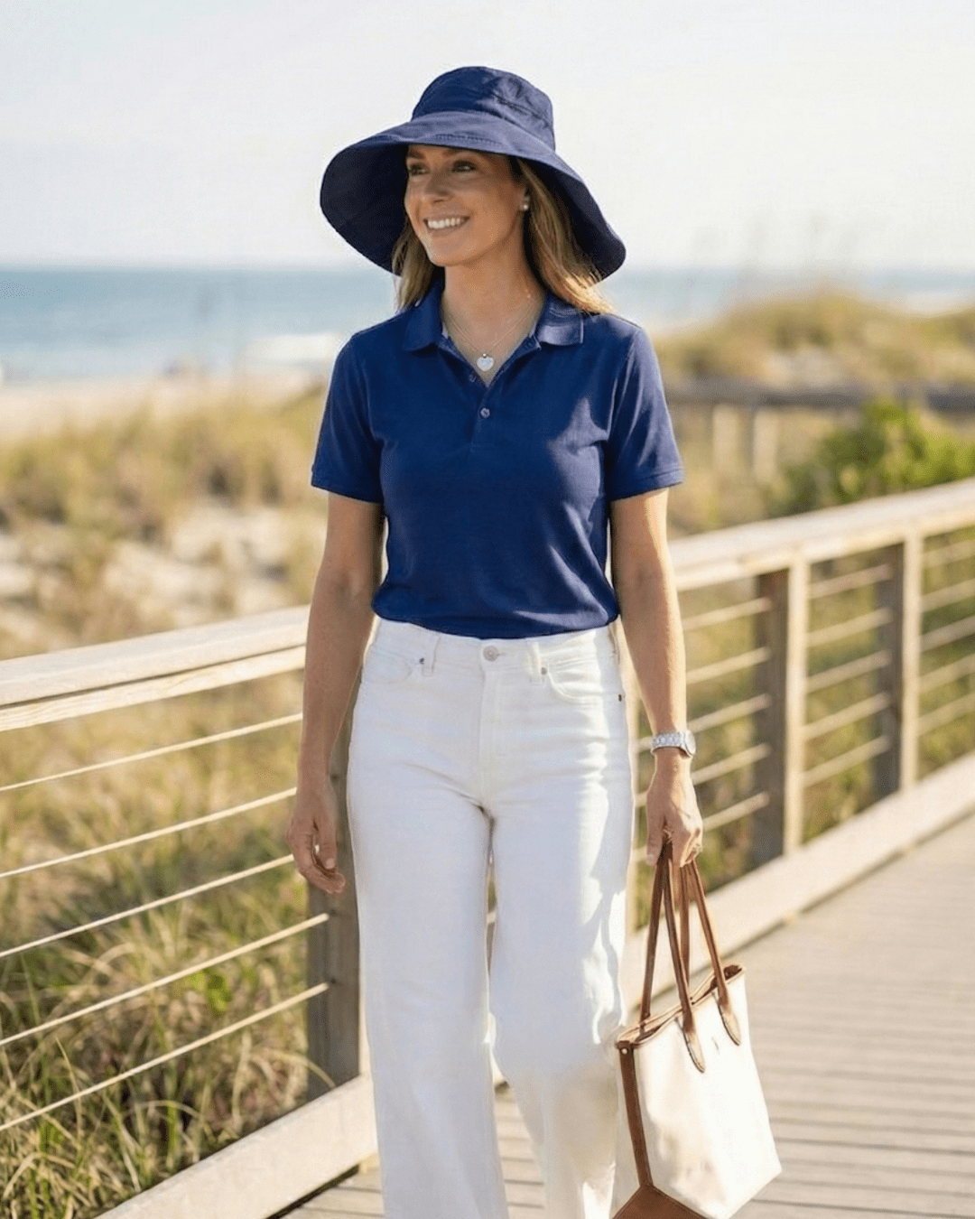 Woman in navy UPF bucket hat and blue UV polo shirt standing on a wooden boardwalk with beach background