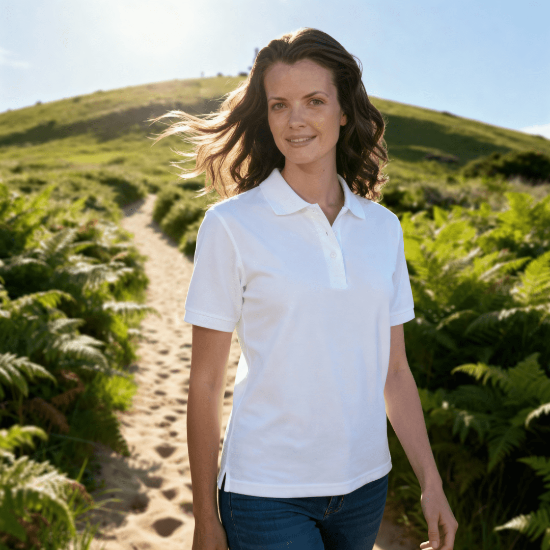 Woman in a white uv golf polo shirt standing on a path with greenery and a clear sky.