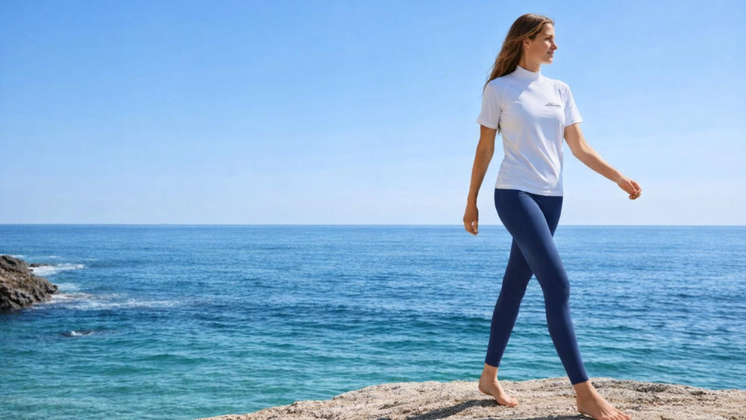 Woman walking on a rocky outcrop by the ocean with a clear blue sky, she is wearing UV protection swim leggings.